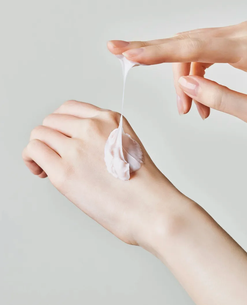 Hand applying a small amount of cream to another hand against a neutral background