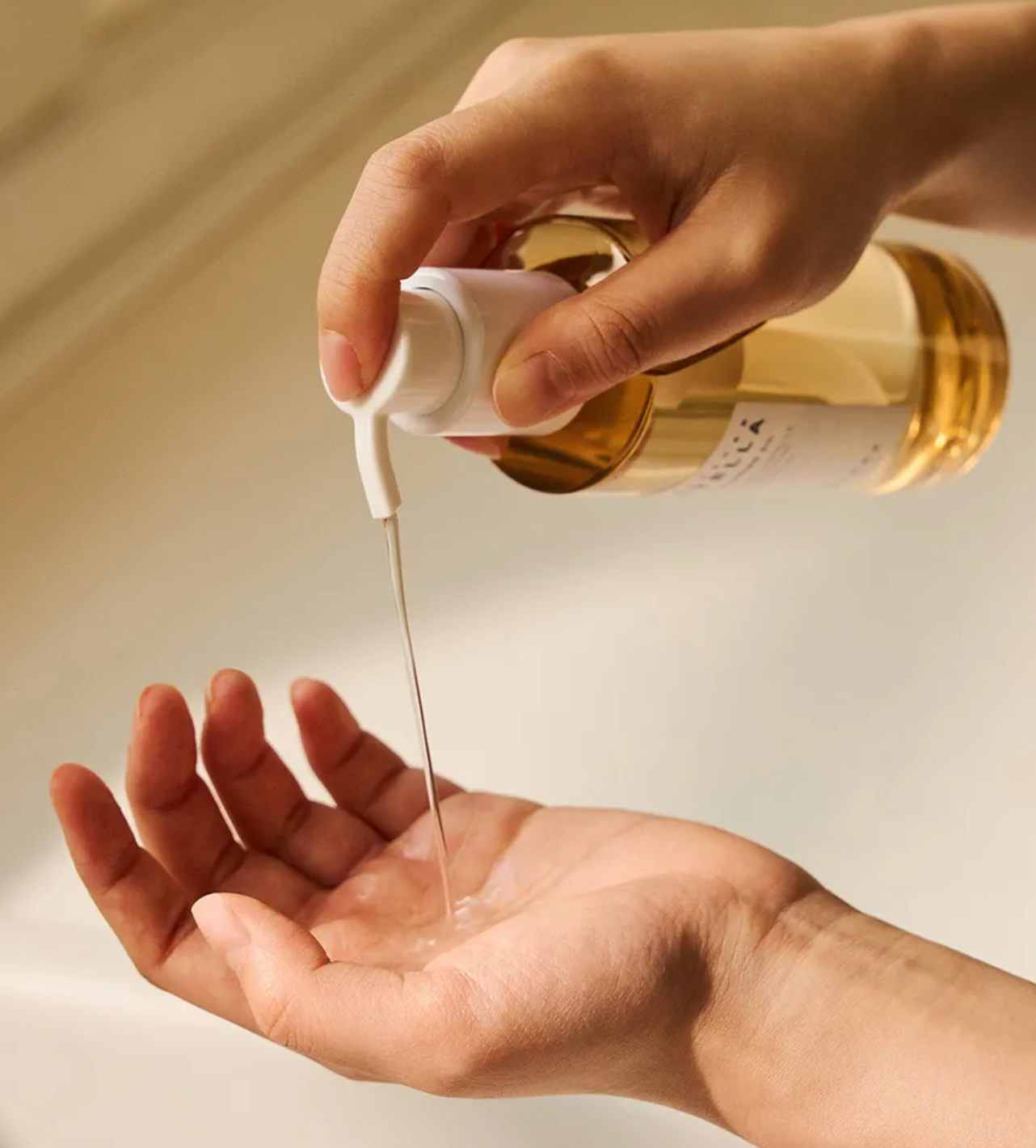 Hand pouring a liquid from a gold bottle into another hand against a neutral background
